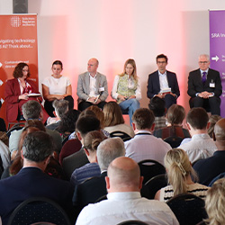 A panel of six people is seated on a stage at a conference. They are facing an audience. The backdrop features text related to navigating regulatory challenges and technology. Audience members are visible in the foreground.